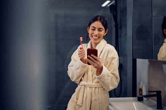 Happy woman reading text message on cell phone while brushing teeth in bathroom.