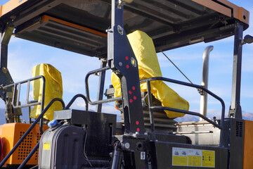 The image features a close-up of a construction vehicle's operator seats, covered in bright yellow protective fabric. The machinery has a black frame and visible controls.