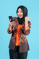Smiling young Asian Muslim woman in batik outer and hijab pointing her thumb while holding a...