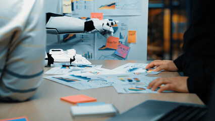 A professional team engages in a business strategy meeting as a robotic arm assists them. They analyze various charts and data, showcasing modern technology in the workspace.Noogenesis