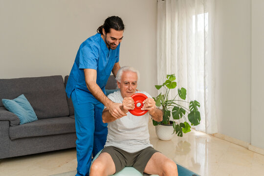 Male Physiotherapist in blue uniform assisting senior man exercising with weights and fitness ball at home