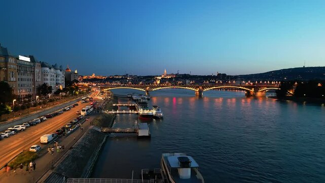 4K drone aerial night video of the Danube riverside in Budapest, Hungary with tourist boats docked along the shore, illuminated streets, and a glowing view of the Margit Bridge under city lights