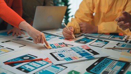 A diverse team engages in a collaborative session, strategizing the design of a mobile application with colorful wireframes and various prototypes laid out on the table. SACTR