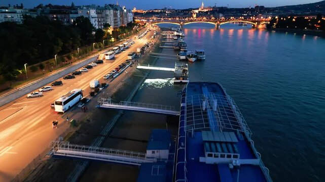 4K drone aerial night video of the Danube riverside in Budapest, Hungary with tourist boats docked along the shore, illuminated streets, and a glowing view of the Margit Bridge under city lights