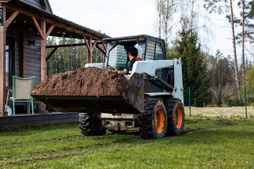 A skid steer loader with orange wheels moves soil on grass near a wooden house with a covered...