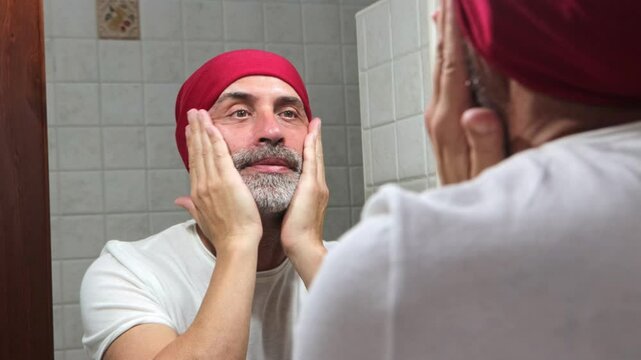 A mature man applying facial cream to his beard in the mirror. Focused on beard care, demonstrating personal hygiene and wellness habits.