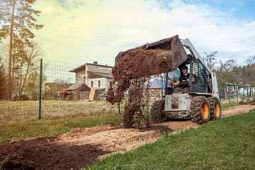 Obraz premium A skid steer loader with orange wheels dumps soil on a pathway near a wooden house with a slanted roof, surrounded by fields and sparse trees.