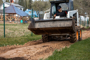 Fototapeta premium A man uses a skid steer loader with a front bucket to level a dirt path. Green wire fence and residential houses with pitched roofs are visible nearby.
