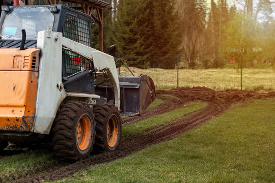 A compact orange and white skid steer loader moves across a grassy field, leaving tire tracks. A wire fence and tall trees are visible in the distance.