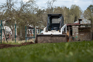 A white skid steer loader with its bucket lowered sits on grass, surrounded by sparse trees, a fenced area, a shed, and a house in a rural setting. © True Pixel Art