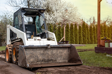 A skid steer loader with a front bucket sits on a dirt path, surrounded by grass, blooming white trees, tall hedges, and a wooden post with flower pots.