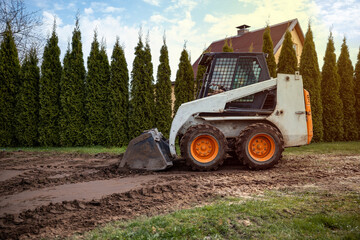 A skid steer loader with orange wheels rests on a dirt path with visible tire tracks. Tall evergreen trees and a house with a sloped roof are nearby. © True Pixel Art