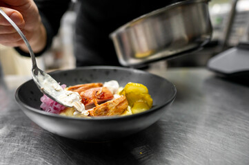 A chef adds a dollop of creamy sauce on a beautifully plated dish featuring chicken, pickles, and garnishes. The attention to detail highlights the culinary artistry in a professional kitchen