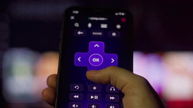 Hand holding smartphone displaying wireless remote control app, pressing button near blurred smart tv screen, representing digital home entertainment connectivity