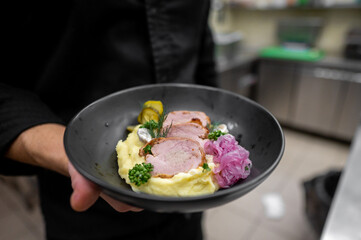 A chef holds a black plate featuring tender slices of meat on a bed of creamy mashed potatoes, garnished with herbs and colorful pickled vegetables, showcasing culinary artistry.