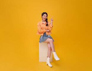 Happy cute Asian teen girl sitting on chair and relax hand pointing finger up isolated on yellow  background. People lifestyle concept.