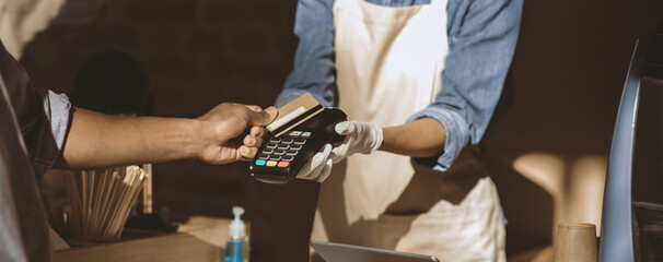 Payment for order and social distance during COVID-19 epidemic. Young african american barista in apron and gloves gives terminal for payment to client in modern cafe, cropped, panorama