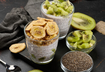 Two glasses of chia seed pudding, one topped with diced kiwi and the other with granola and banana chips, are on a dark surface with ingredients
