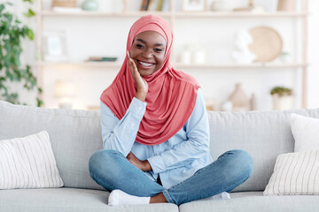 Obraz premium Traditional Black Muslim Woman In Hijab Posing On Couch At Home, Smiling At Camera, Religious Islamic Lady Relaxing On Sofa In Living Room, Resting Chin On Hand, Enjoying Domestic Weekend, Copy Space