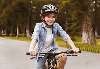 Portrait of cute curly little boy riding a bicycle in public park and smiling, blurred background