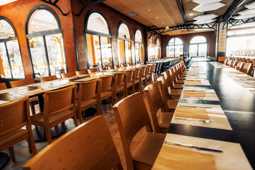 Spacious empty restaurant interior with long neatly set dining tables, wooden chairs, arched windows, and warm ambient lighting in a modern elegant design; wide-angle indoor shot