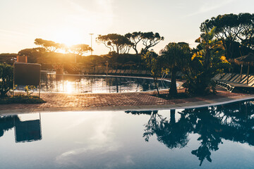 Tropical resort pool at sunrise in Punta Umbria, Spain, with palm trees, sun loungers, and still water reflections under a warm light, evoking a tranquil summer escape; wide-angle shot of golden hour