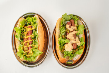 Flat lay of two hot dogs on oval stainless steel plates, garnished with lettuce, pickles, mustard, jalapenos, and creamy dressing, on white background