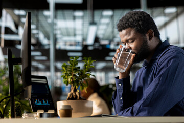 African american business employee drinking a glass of water and hydrating at the corporation, taking care of his body. Male worker takes a break from work in order to hydrate, wellbeing.