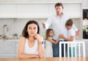 Offended mother sitting at the kitchen table, her husband, son and little daughter standing behind...