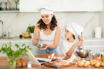Happy mother and daughter are baking salmon in the kitchen while looking at the recipe on laptop