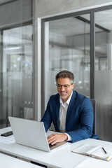 Fototapeta premium Mature Latin business man ceo manager using laptop computer in modern office. Smiling senior businessman entrepreneur sitting at desk working on financial project, looking thinking at screen. Vertical