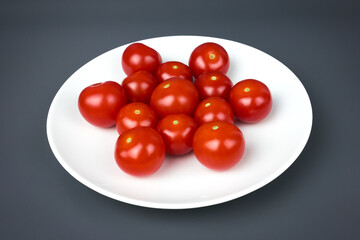 Small red ripe cherry tomatoes on a white plate, gray background, close-up. Vegetables for dietary healthy eating and weight loss.