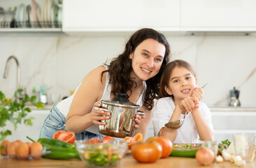 Mom and little daughter are standing at kitchen table with dishes in their hands. Girl helped her mother cook lunch, learned to perform culinary masterpieces.
