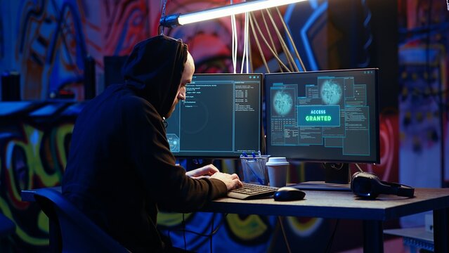 Hooded hacker in graffiti painted hideaway targeting vulnerable unpatched connections, having burst of joy after getting access granted past security and stealing valuable sensitive data