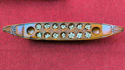 Top-down view of a traditional Congklak board game with seeds placed in carved wooden pits, set against a red textured background.