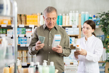 Senior man buys emollient cream at pharmacy and compare with other medicine, carefully reads patient information, checks composition of drug. Girl pharmacist offer to buy similar drug.
