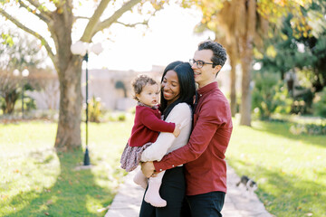 Fototapeta premium Smiling dad hugging mom from behind with a little girl in her arms in the sunny park