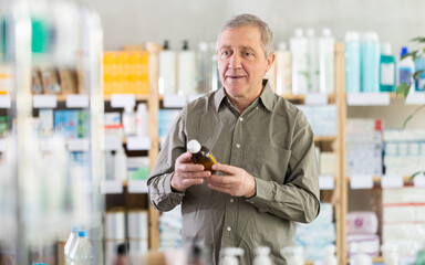 Elderly man in casual olive shirt carefully examining brown medicine bottle in pharmacy, checking...