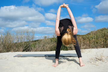 Woman practicing yoga outdoors on sandy surface, performing forward bend with clasped hands behind back, embracing balance, flexibility, and mindfulness under bright sky and nature
