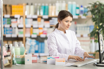 Positive skilled young female pharmacy worker in white coat using desktop computer to update product quantities and manage inventory during routine stock control