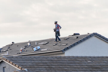 A roofer in a safety harness walks across the pitched roof of a newly constructed home, where loose tiles, vents, and equipment are scattered during installation © Eduardo Barraza