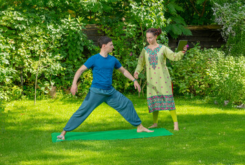 female instructor in green sari doing yoga with male student in garden on grass