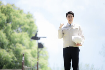 新緑の屋外で作業着姿でヘルメットを持つ上半身　施工管理や現場などのイメージ　カメラ目線でガッツポーズ