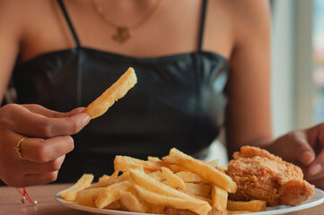 Woman picking up golden french fry from plate, crispy fried chicken nearby, savoring casual...