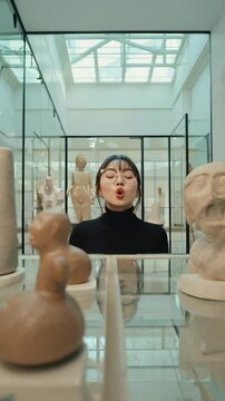 Young adult female wearing glasses making a kissy face at museum sculptures in glass cases with bright daylight streaming in.