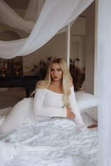 Fototapeta premium Front view of a young blonde woman in an off-shoulder white dress, lying on a white bed under a sheer canopy, looking at camera. Concept lifestyle, beauty, bedroom decor.