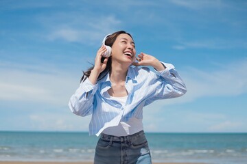 A joyful young Asian woman listening to music with headphones at the beach, smiling brightly under the clear blue sky.enjoying and relaxing summer atmosphere concept.