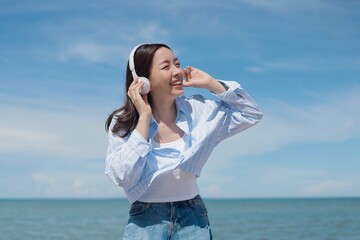 A joyful young Asian woman listening to music with headphones at the beach, smiling brightly under the clear blue sky.enjoying and relaxing summer atmosphere concept.