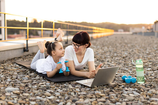 Young mother and daughter using laptop for online exercise session on rooftop, enjoying quality bonding and fitness. Family spends time healthily while performing fun
