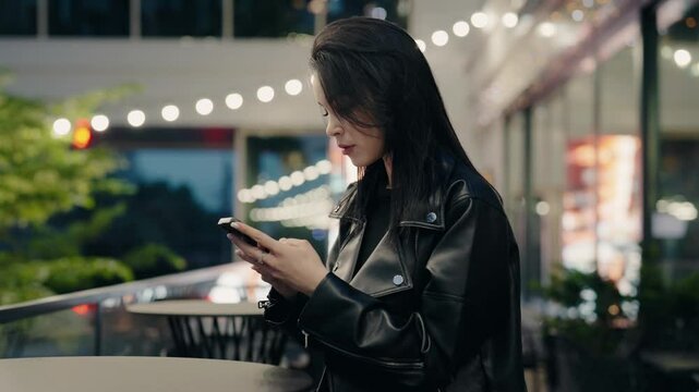 Young woman in leather jacket using smartphone while standing outdoors under string lights at night, surrounded by a modern city ambiance. Woman in Leather Jacket Texting Outdoors at Night